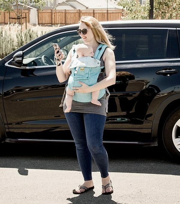 Looking at YourMechanic app Mom standing outside with baby in baby carrier looking at YourMechanic app