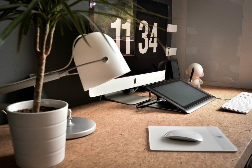 desk with computer, lamp and plant on it with the time 11:34 and a wireless mouse
