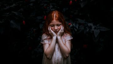 redhaired girl with hands over her cheeks in front of dark background- looks worried