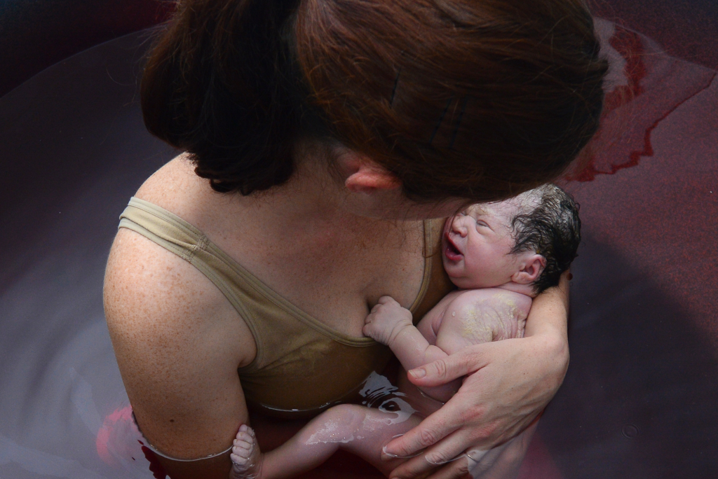 postpartum mom holds newborn in bloody birth tub